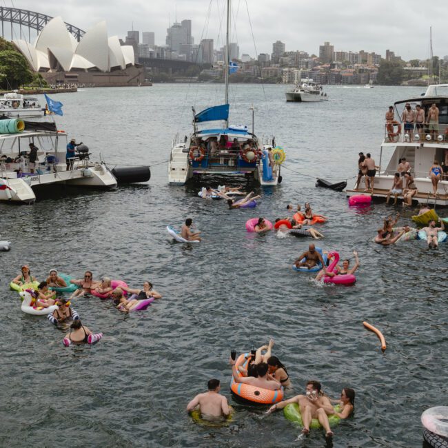 People enjoying a summer day in the water near anchored boats, using inflatables in a bay. The Sydney Opera House and Harbour Bridge are visible in the background under a cloudy sky.