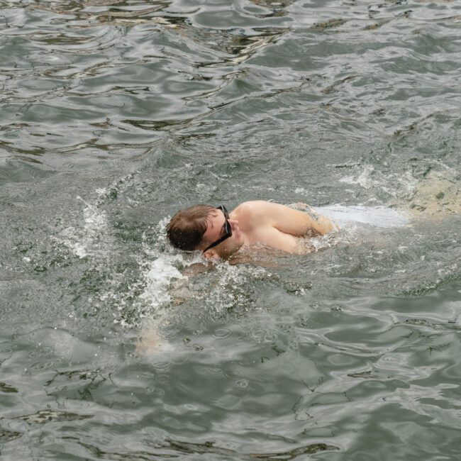 A person swimming freestyle in a body of water. They are wearing goggles and are partially submerged, creating splashes with their strokes. The water appears rippled and slightly turbulent around them.