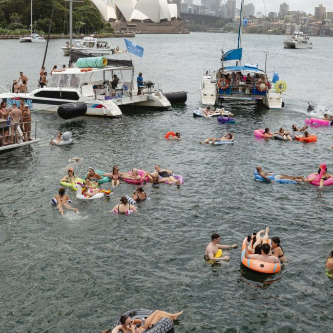 People enjoy a floating party in Sydney Harbour, surrounded by boats and using inflatable rafts and pool toys. The Sydney Opera House and Harbour Bridge are visible in the background under cloudy skies.