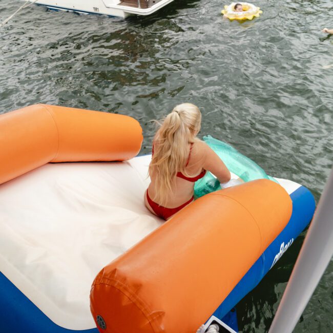 A person in a red swimsuit is sitting on an orange and white inflatable float on a lake. Nearby, two more people are enjoying the water on inflatable rings. A boat is partially visible in the background.