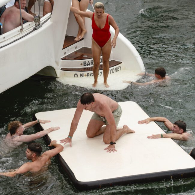 A group of people enjoying the water near a boat. One person in a red swimsuit stands on the boat's edge, while another kneels on a floating mat. Others swim around nearby, with a large blue umbrella partially visible in the foreground.