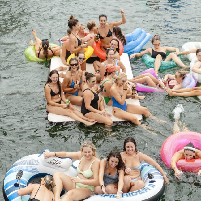 A group of people, mostly young women, are lounging and socializing on colorful inflatable rafts and pool floats in the water, enjoying a sunny day. The scene is lively with smiles and drinks, featuring various float designs like a unicorn and paw prints.