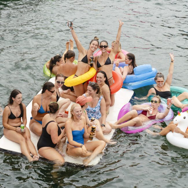 A group of people in swimsuits enjoying a sunny day on a lake, sitting on inflatable rafts and pool floats. Some hold drinks, while others wave and smile. The water is calm, and more people are seen floating in the background.