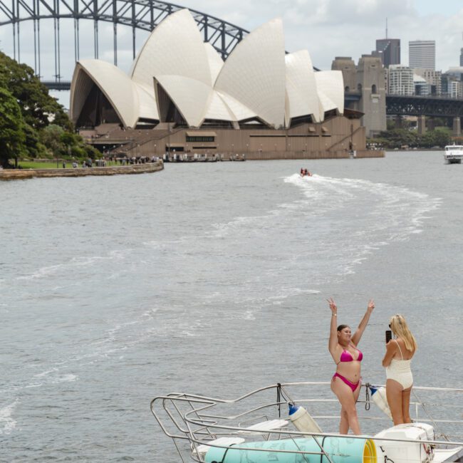 Two people stand on a boat in Sydney Harbour, with one posing energetically. The Sydney Opera House and Harbour Bridge are visible in the background. The sky is partly cloudy. The boat has a rolled-up mat on the deck.