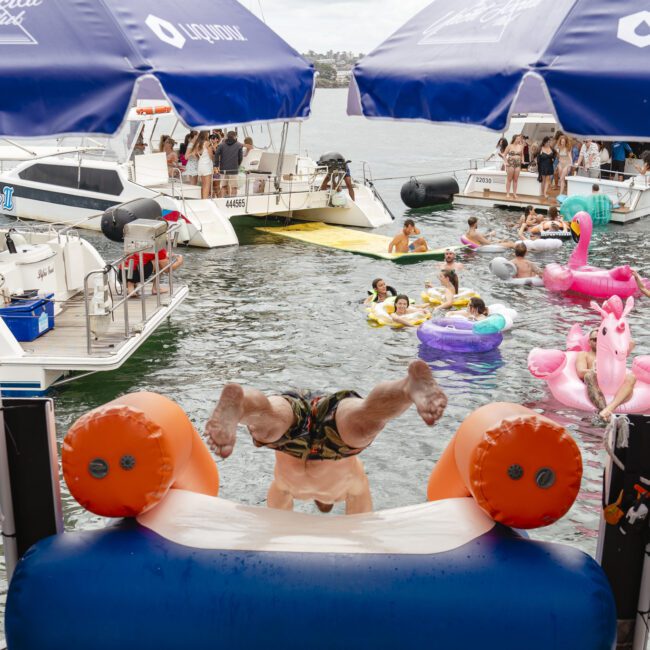 A person is diving headfirst from an inflatable platform into a water area surrounded by boats and people on colorful inflatables. Lifeguards in yellow shirts are overseeing the activity, and a person is taking a photo with a smartphone.