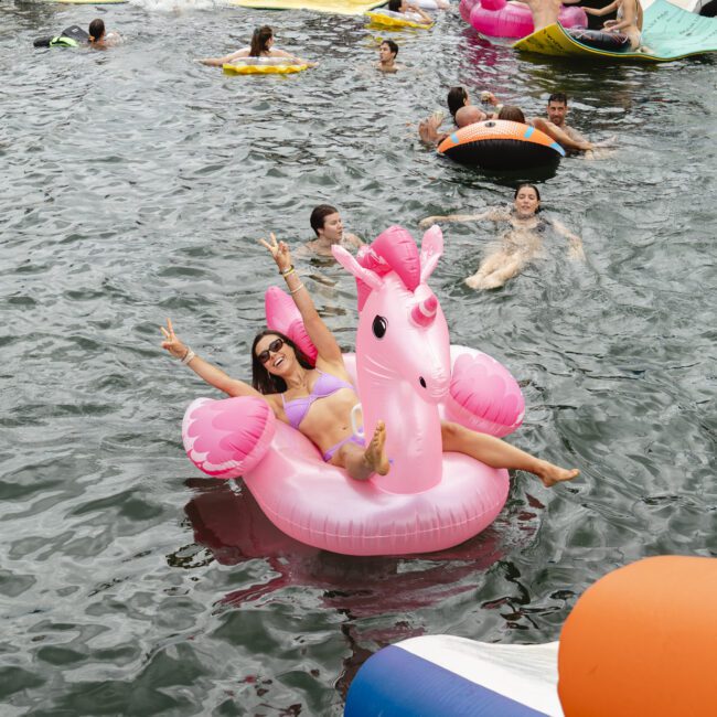 People are having fun swimming and relaxing on inflatable floaties in a body of water near boats. A person is posing with peace signs on a large pink unicorn float. Others are on round inflatables nearby.