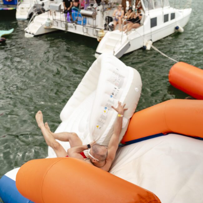 A person in a swimsuit is sliding off an inflatable orange and white float into the water. Boats with people relaxing are visible in the background, and a large flamingo float is floating nearby.