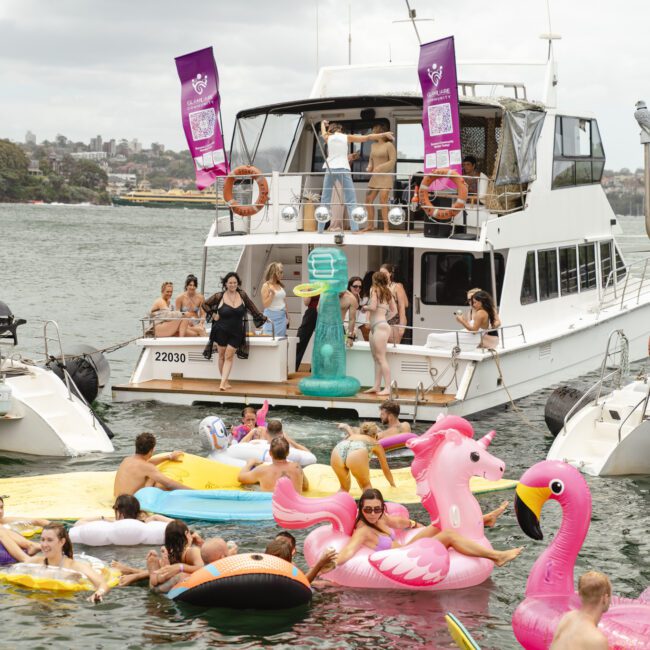 A lively scene on the water with people enjoying a party on a boat and various inflatable floats. The bright floats include flamingos, unicorns, and other shapes. Attendees are in swimsuits, with a backdrop of cloudy skies and distant shoreline.