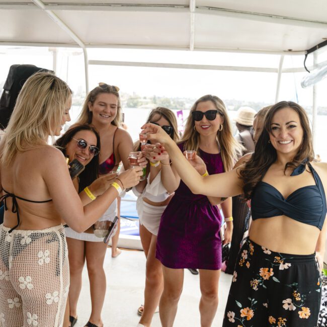 A group of women on a boat, some wearing swimsuits and others in casual clothing. They are raising glasses in a toast, smiling and enjoying the sunny day. The background shows a body of water and a partly cloudy sky.