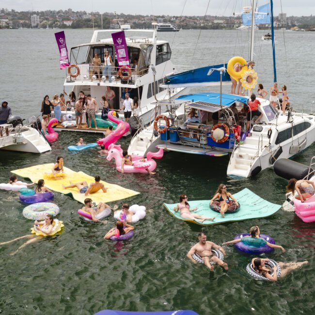 People enjoying a party on yachts and inflatables in a sunny harbor. The scene includes various colorful floaties, such as flamingos and unicorns, with attendees relaxing and socializing on boats and in the water.