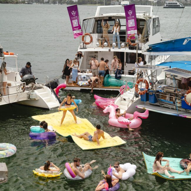 A group of people enjoying a party on boats and inflatables in the water. Some are sitting on colorful floats shaped like flamingos and mattresses, while others are socializing on the boats. The scene is lively and festive.