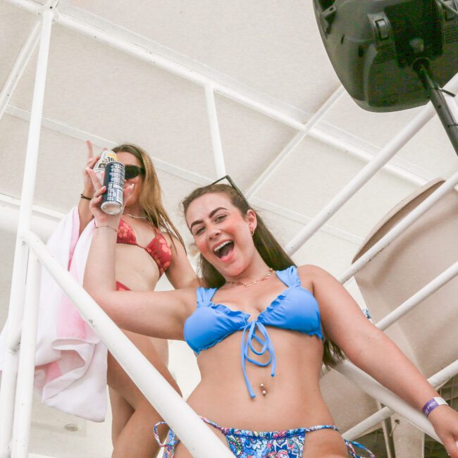 Two women in swimsuits are on a boat's staircase. The woman in the foreground wears a blue bikini, holding a drink can, and smiles at the camera. The woman behind wears a red bikini and holds a towel and a peace sign. A speaker is mounted on the railing.