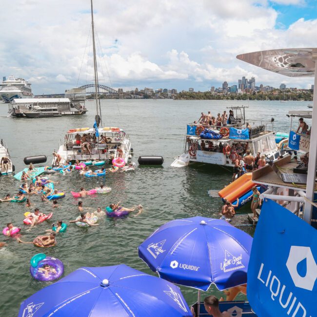 A lively scene of a floating party with people enjoying the water on colorful inflatables. Several boats are anchored nearby, and a DJ stands on one boat playing music. The city skyline and a bridge are visible in the background under a partly cloudy sky.