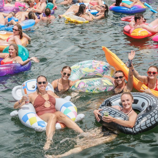 A group of people are floating on colorful inflatable tubes in a body of water, smiling and waving. The scene is lively and joyful, with many more people in the background also enjoying the water on various inflatables.