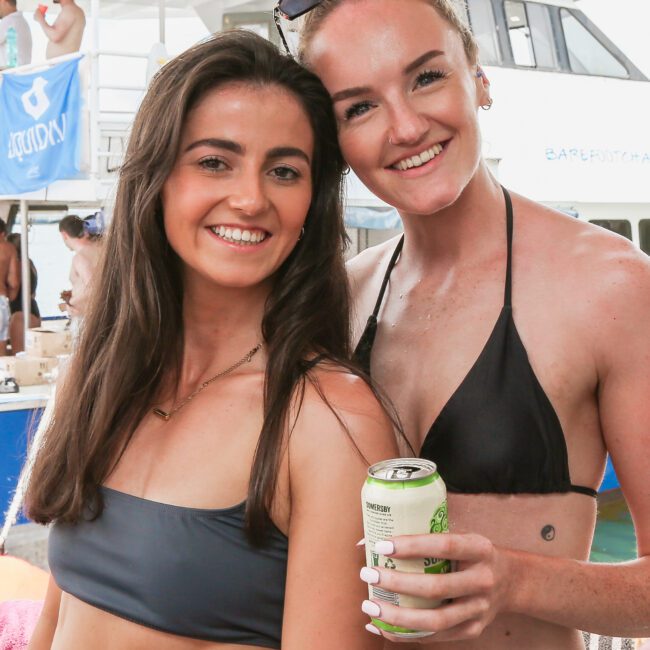 Two women in swimsuits smile for a photo on a boat. One woman holds a can. Other people are in the background near the boat's deck. They appear to be enjoying a sunny day on the water.