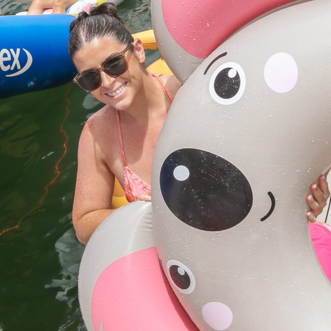 A smiling woman in sunglasses and a pink swimsuit is in the water holding a koala-shaped pool float. Other colorful inflatable floats and people are around her in the background.