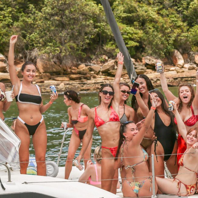 A group of people in swimsuits celebrating on a boat with drinks. They are smiling and raising their arms, surrounded by water and lush greenery.