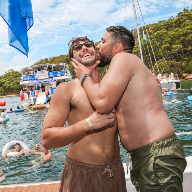 Two shirtless men are on a boat. One man is giving the other a playful kiss on the cheek. Behind them, people are swimming and enjoying a party on several boats, with inflatables floating in the water. The scene is sunny with trees in the background.