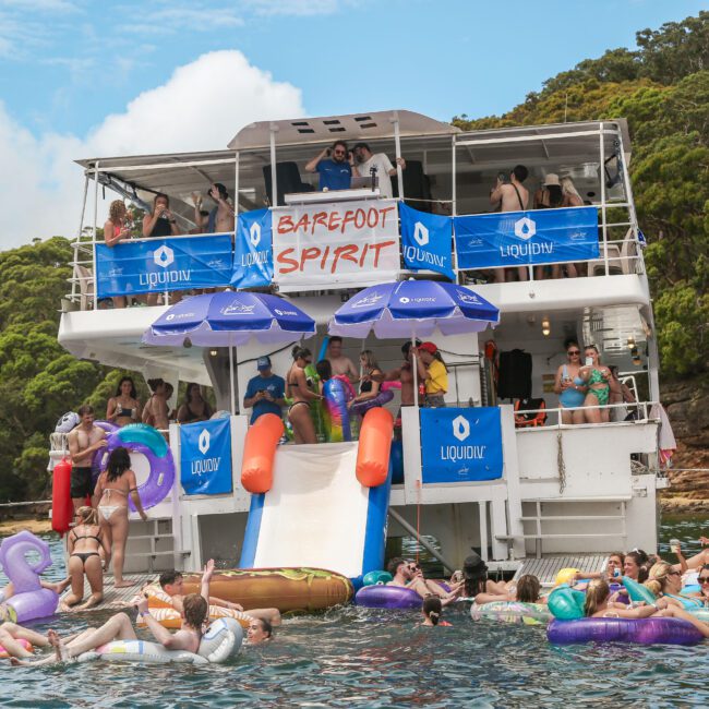 A large party boat with "Barefoot Spirit" written on it is docked near wooded shorelines. People are enjoying themselves on inflatable floats in the water, while others gather on the boat under umbrellas. The atmosphere is lively and festive.