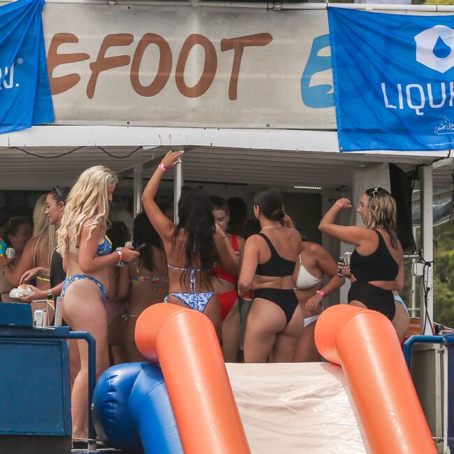 A group of people in swimsuits enjoying themselves on a boat with a slide. The boat is decorated with blue banners that say "Liquid" and is anchored in a sunny, outdoor setting.