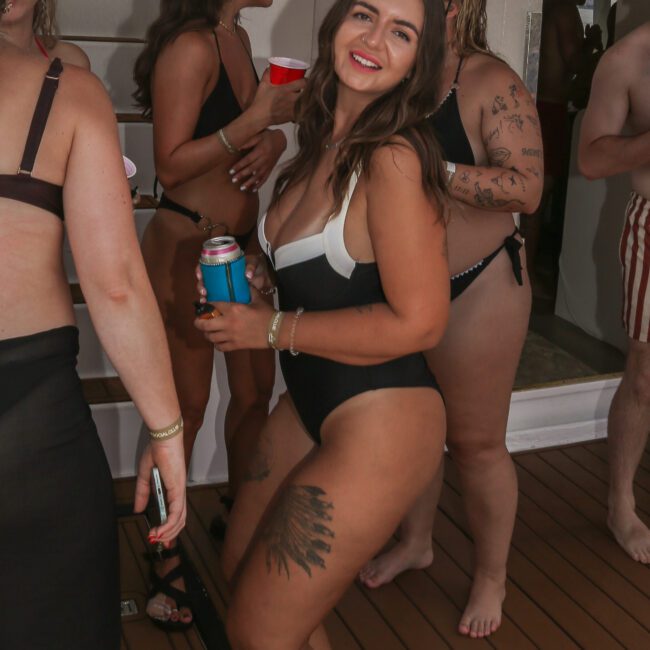 Group of people in swimwear enjoying a party on a boat. A woman in the foreground smiles while holding a drink. Other partygoers are mingling in the background.