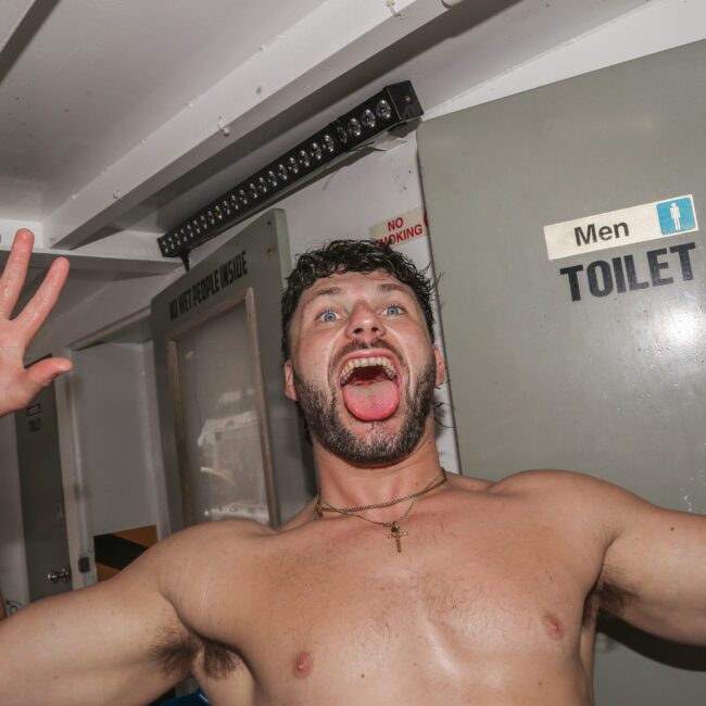 A shirtless man with curly hair stands in front of a door labeled "Men Toilet," holding a can in one hand and raising the other with fingers spread wide. He has a playful expression with his mouth open and tongue out.