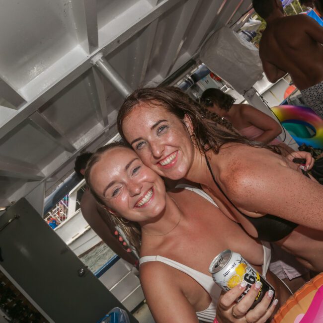 Two women smiling and posing together on a boat. One is holding a drink can. Both are dressed in swimwear. In the background, other people are enjoying the boat party, some with colorful inflatable pool toys.