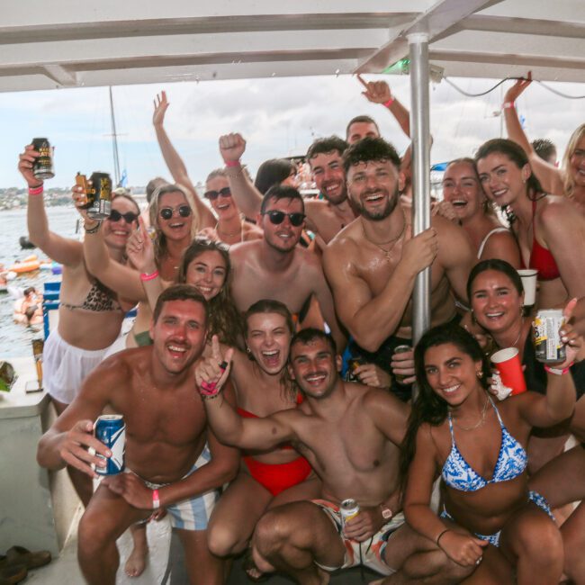 A group of smiling people in swimsuits and swim trunks pose together on a boat, holding drinks and showing excitement. The background features a scenic view of water and land under a cloudy sky.