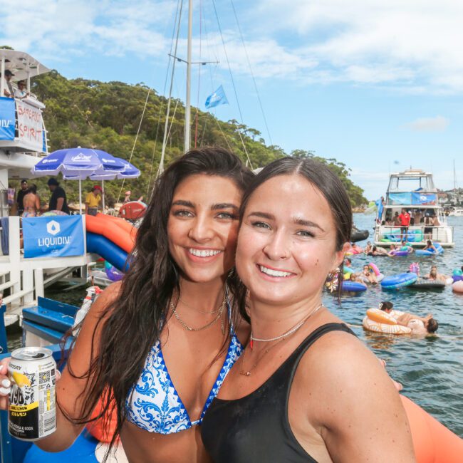 Two women in swimsuits smile for a photo on a boat. One holds a can in her hand. Behind them, people are swimming and enjoying the sunny day. A blue sky and trees are visible in the background.