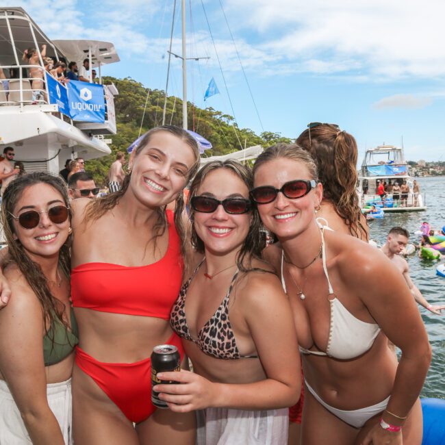 A group of four smiling women in swimsuits pose together on a boat. In the background, people are socializing on another boat and in the water. The scene is lively, with clear skies and inflatable floats around.