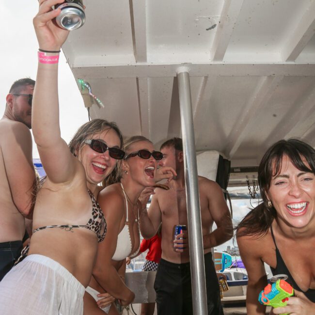 A group of people in swimwear enjoying a lively party on a boat. They are smiling, holding drinks, and some are wearing sunglasses. The atmosphere appears festive and energetic.