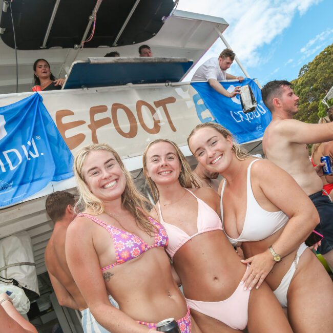 A group of people in swimsuits are enjoying a sunny day on a boat. Three women are smiling at the camera in the foreground. A "Liquid IV" banner is visible in the background. Trees and a partly cloudy sky are in the distance.