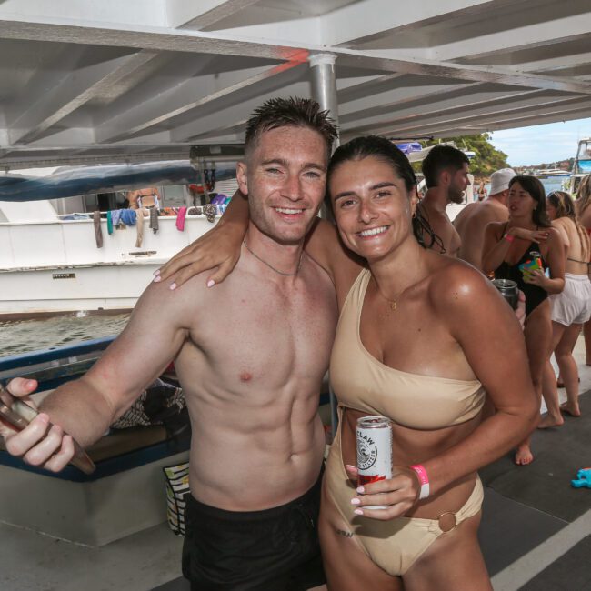 A man and a woman smiling on a boat, with the man holding a phone. The woman is wearing a beige bikini and holding a can. People are visible in the background, enjoying the boat party.