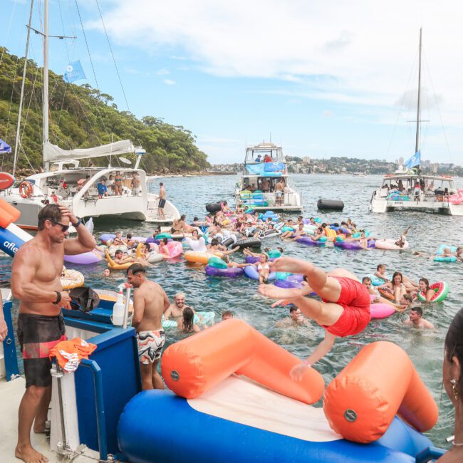 People are enjoying a lively boat party. A person dives into the water from a float. Several colorful inflatables and boats are in the water, surrounded by trees and a clear sky in the background.