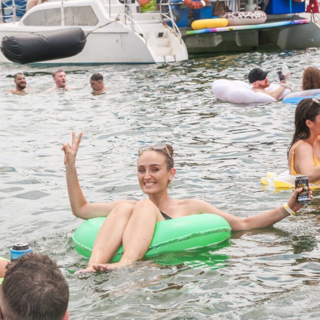 A group of people relax on inflatable floats in a body of water, surrounded by boats. One woman in the foreground on a green float smiles and gestures with her hand. Others enjoy floating on various colored inflatables under a cloudy sky.