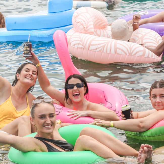 A group of people enjoying a sunny day on inflatable floaties in the water. They are smiling and holding drinks, with various colorful floats, including a pink flamingo and a green circle. The atmosphere is lively and fun.