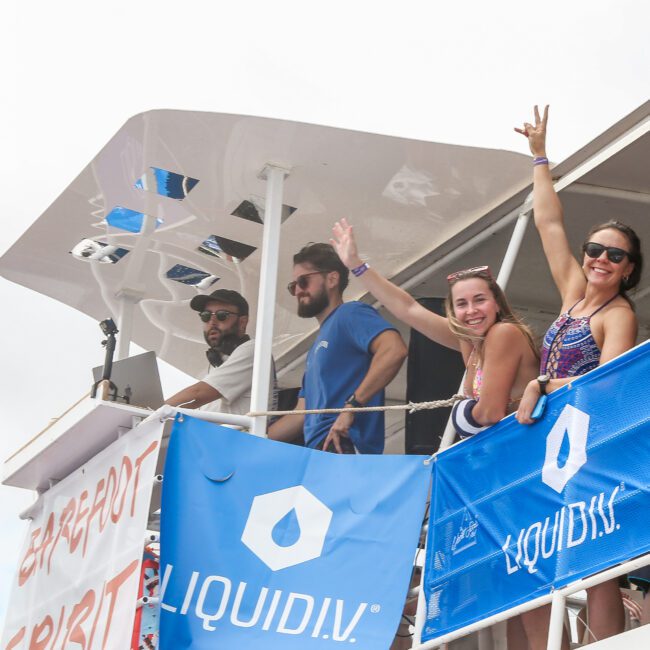 A group of people stands on a boat deck with blue Liquid I.V. banners. Two women are smiling and waving, while two men, one wearing sunglasses, watch. They appear to be enjoying a sunny day on the water.