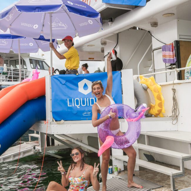 Two women in swimsuits pose on a boat's deck. One stands holding a purple inflatable ring, the other sits flashing peace signs. A man under a purple umbrella stands on the boat above, with a "Liquid" banner visible.