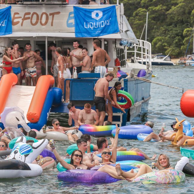 A lively party scene with people on a boat and others enjoying inflatable floats in the water. The boat has banners and slides. Various colorful floats, including unicorns and swans, are in use. The background features trees and moored boats.