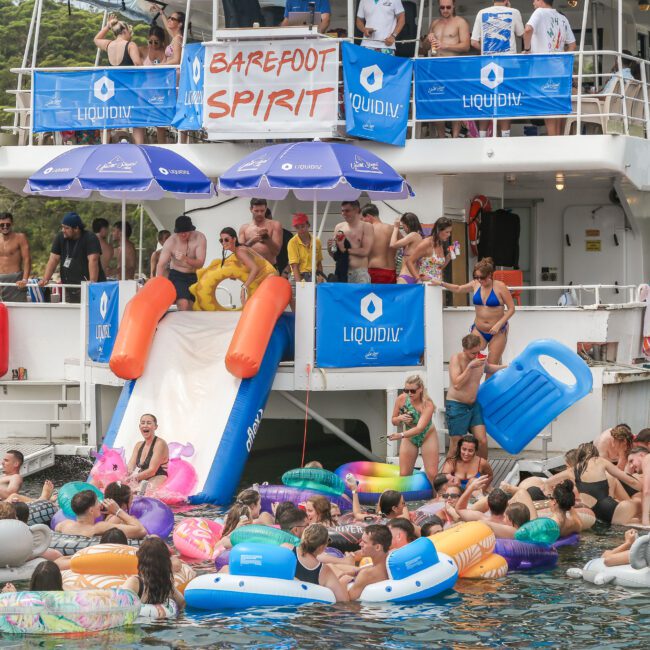 A lively boat party with people enjoying a large inflatable slide, surrounded by colorful pool floats in the water. Signs with "Barefoot Spirit" and "LiquidII" are visible. The background features lush greenery.