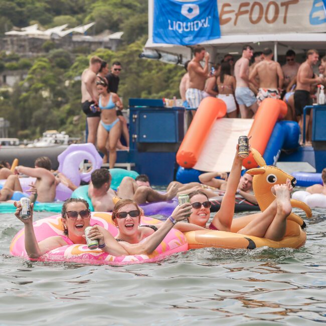 People enjoying a party on water, sitting on inflatable floats with drinks in hand. The scene includes a boat filled with more party-goers and lush greenery in the background.