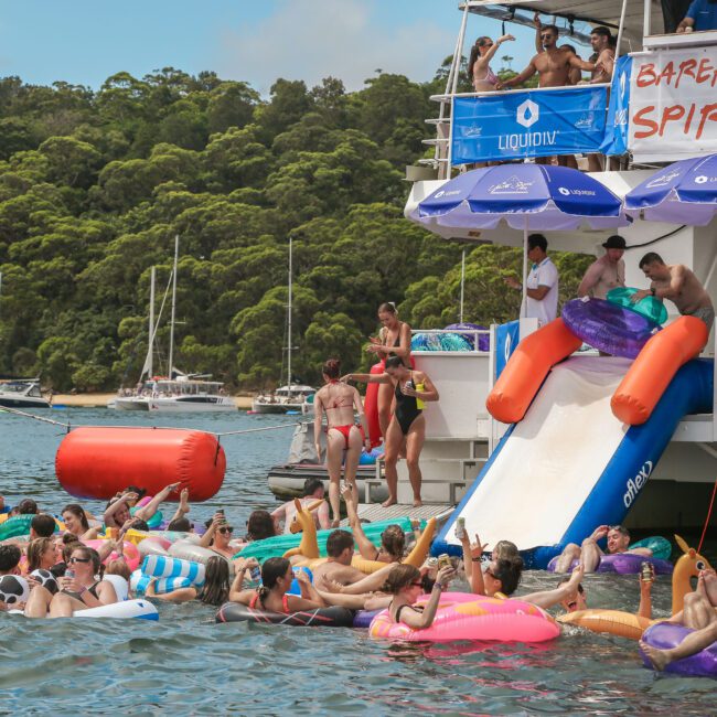 A lively boat party featuring people in swimsuits enjoying a sunny day. Some are on inflatable floats in the water, while others use a slide from the boat. Trees and boats in the background add to the festive atmosphere.