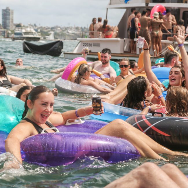 A lively group of people enjoying a water party, lounging on colorful inflatables in a bay. They're smiling, holding drinks, and surrounded by floating pool toys. A boat and distant cityscape are in the background under a partially cloudy sky.