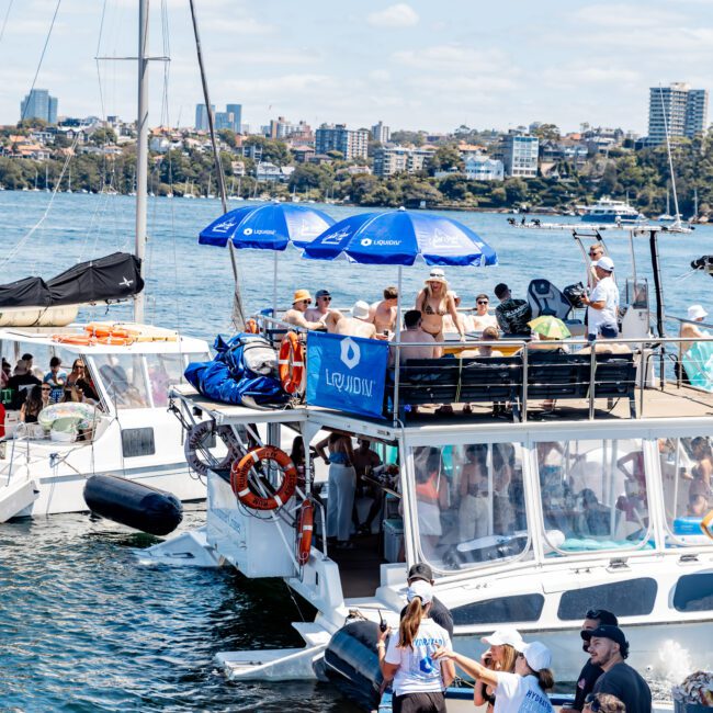 People are enjoying a sunny day on boats at a lively waterfront gathering. Some are on a large boat with blue sun umbrellas, while others relax on inflatable floats in the water. The city skyline is visible in the background.