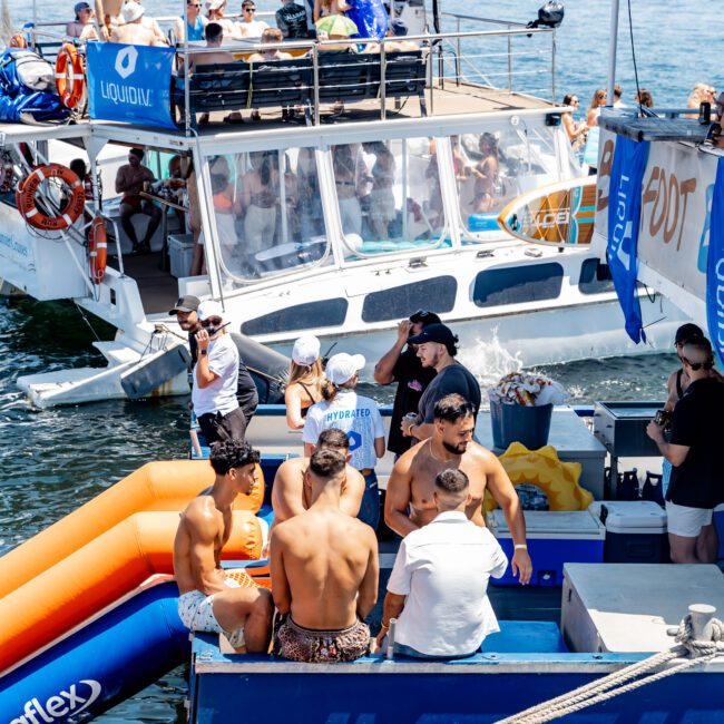 A group of people socializing on a boat anchored near the shore. Some are seated while others stand on deck. An inflatable orange slide is attached to the side of the boat. City buildings are visible in the background under a clear sky.