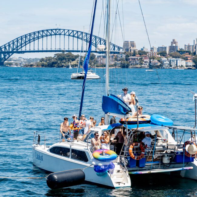 A group of people enjoying a sunny day on a sailboat with rubber boats and life rings. In the background, a large bridge and cityscape are visible across the water.