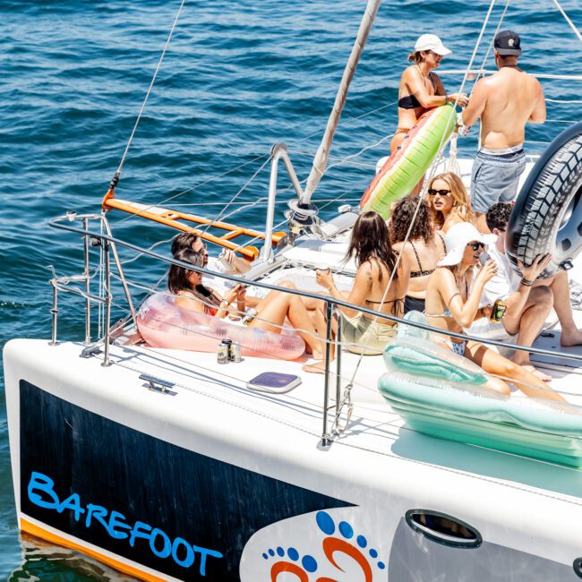 A group of people lounging on the deck of a sailboat named "Barefoot." They are sitting on inflatable cushions, enjoying the sunny weather on the water. One person is standing at the back, and others are reclining and conversing.