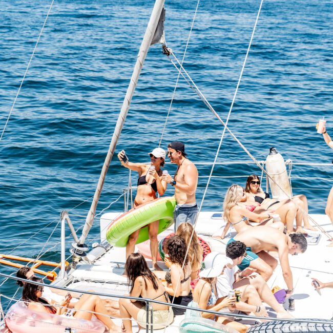 A group of people is enjoying a sunny day on a yacht. Some are sitting or lying on the deck, and two individuals are taking a selfie. Inflatable pool floats and the city skyline are visible in the background.