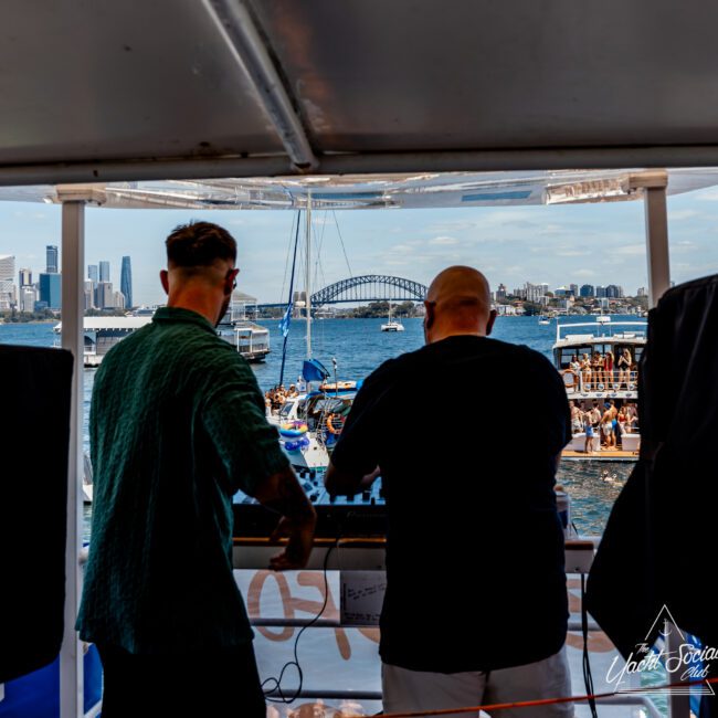 Two DJs perform on a boat with a crowd in the background. The Sydney skyline and Harbour Bridge are visible against a partly cloudy sky. The scene captures a lively waterfront atmosphere.