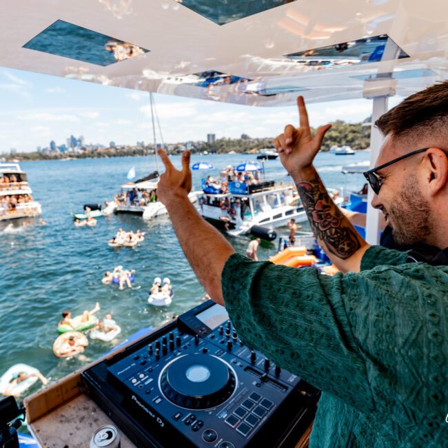 A DJ wearing sunglasses and a green shirt is playing music on a boat. He is pointing upwards with both hands. In the background, people on boats and inflatables enjoy the water on a sunny day.